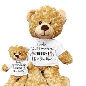 May include: Two brown teddy bears wearing white t-shirts with the text "Cindy. You're winning the fight. I love you. Mom" and pink ribbon graphics. The larger bear is standing and the smaller bear is sitting.