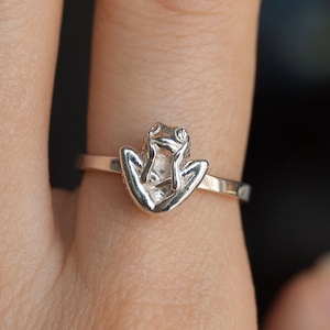 Puede incluir: Un anillo de plata con un diseño de rana. La rana está sentada en la banda del anillo con las patas extendidas.