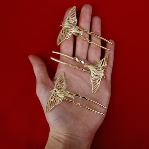 May include: Three gold butterfly hair clips with intricate designs. The clips are held in a hand against a red background.