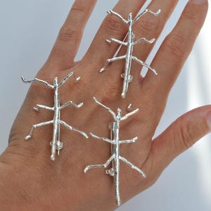 May include: Three silver-colored stick insect brooches. Each brooch is designed to resemble a twig with multiple branches. The brooches are displayed on a light-colored background.