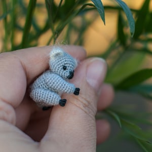 May include: A small grey crocheted koala bear is being held in a person's hand. The koala is sitting up with its arms and legs outstretched. The background is a blurry image of green leaves.