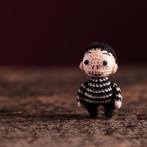 Puede incluir: Una pequeña muñeca de crochet con cabello negro, una camisa a rayas blancas y negras y una expresión facial neutra. La muñeca está de pie sobre una superficie de madera.