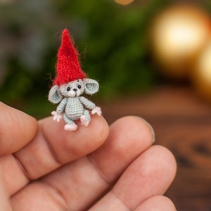 May include: A small crocheted mouse wearing a red Santa hat. The mouse is sitting on a person's finger.