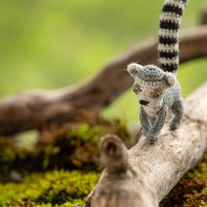 Könnte beinhalten: Ein kleines, grau-weißes Häkel-Lemuren-Spielzeug mit schwarz-weiß gestreiftem Schwanz sitzt auf einem Ast mit grünem Moos im Hintergrund.