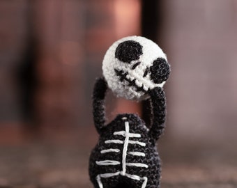 Puede incluir: Un juguete de esqueleto de crochet negro y blanco sosteniendo una calavera de crochet blanca con ojos negros y una boca cosida. El juguete está de pie sobre una superficie de madera.