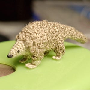 Può includere: Piccola statuina di pangolino fatta a mano all'uncinetto, in filato beige. Il pangolino ha un aspetto strutturato e squamoso, con naso e occhi neri. È posizionato su una superficie verde chiaro. Un oggetto artigianale unico.
