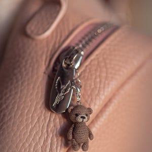 Könnte beinhalten: Nahaufnahme einer rosa Ledertasche mit silbernem Reißverschluss und einem kleinen, gehäkelten Teddybären-Anhänger in Braun. Der Anhänger ist mit einer kleinen Kette am Reißverschluss befestigt. Die Tasche hat eine strukturierte Oberfläche.