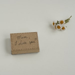 May include: A small brown box with the words "Mom, I love you" written on it. A small bouquet of white crocheted flowers is on a white surface.