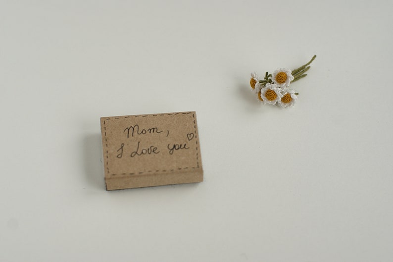 May include: A small brown box with the words "Mom, I love you" written on it. A small bouquet of white crocheted flowers is on a white surface.