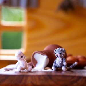 May include: A wooden table displays two small, crocheted cat figurines, one white and one gray, near a brown overturned pitcher with spilled white liquid. The scene evokes a playful, miniature world.