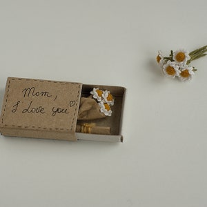 May include: A small matchbox with a handwritten message that says "Mom, I love you" and a small bouquet of crocheted white flowers inside.