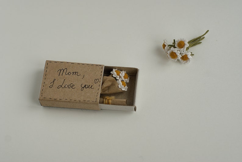 May include: A small matchbox with a handwritten message that says "Mom, I love you" and a small bouquet of crocheted white flowers inside.