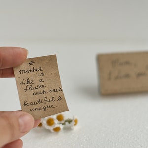 May include: A small brown card with the handwritten message "A mother is like a flower, each one is beautiful & unique." The card is held in a hand with a small bouquet of white flowers in the foreground.