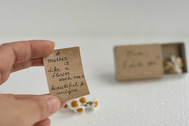 May include: A small brown card with the handwritten message "A mother is like a flower, each one is beautiful & unique." The card is held in a hand with a small bouquet of white flowers in the foreground.