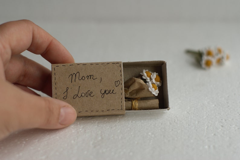 May include: A small brown box with a handwritten message that says "Mom, I love you" and a small bouquet of crocheted white flowers inside.