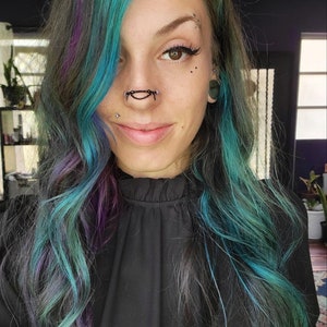 May include: A person with teal, purple, and gray hair. They are wearing a black top and have multiple facial piercings, including a nose chain. The person is smiling at the camera.