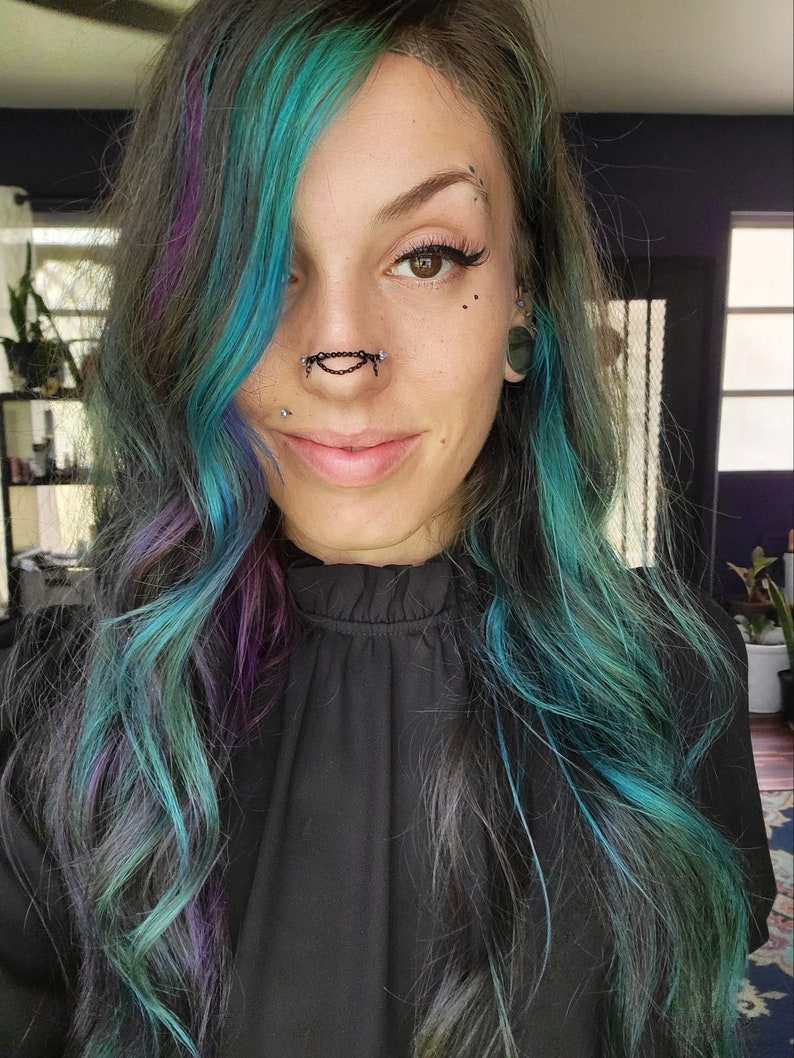 May include: A person with teal, purple, and gray hair. They are wearing a black top and have multiple facial piercings, including a nose chain. The person is smiling at the camera.