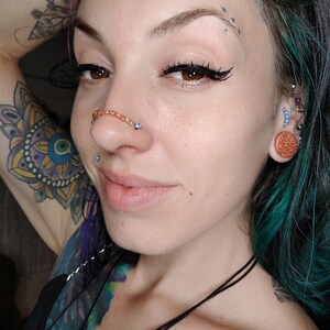 K&ouml;nnte beinhalten: Eine Frau mit t&uuml;rkisfarbenem Haar und einem Nasenring. Sie hat ein Tattoo am Arm und eine Halskette.