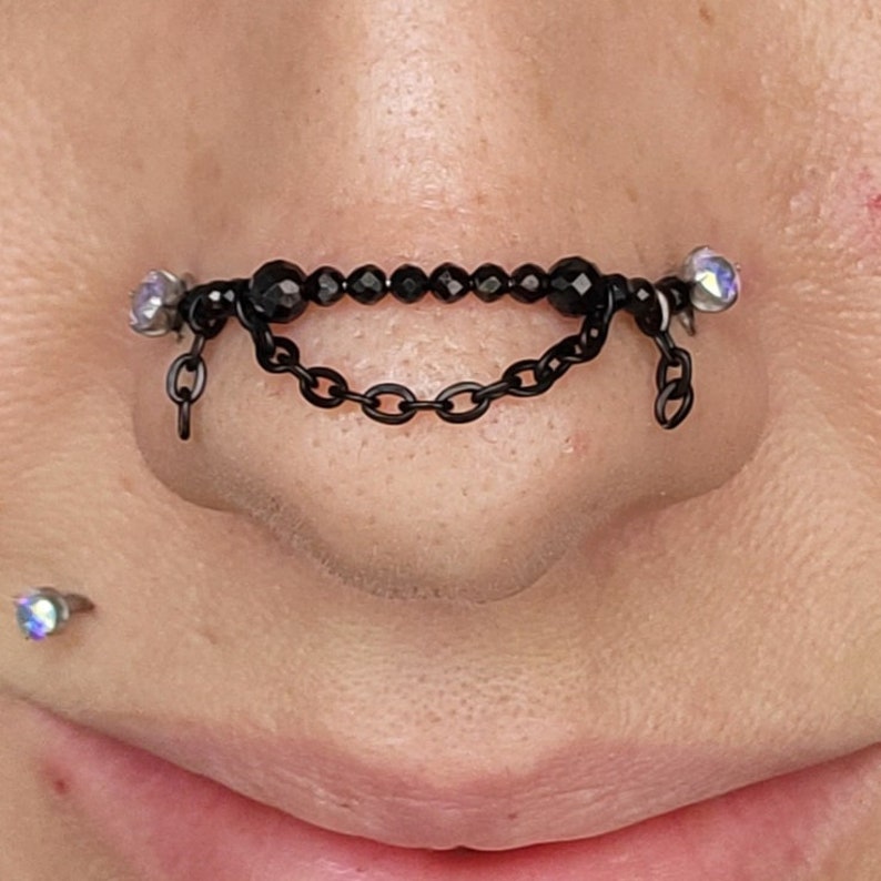 May include: Black nose jewelry featuring a chain and beaded design. The jewelry has a chain that dangles below the nose. The jewelry is adorned with small, iridescent gems on each end.