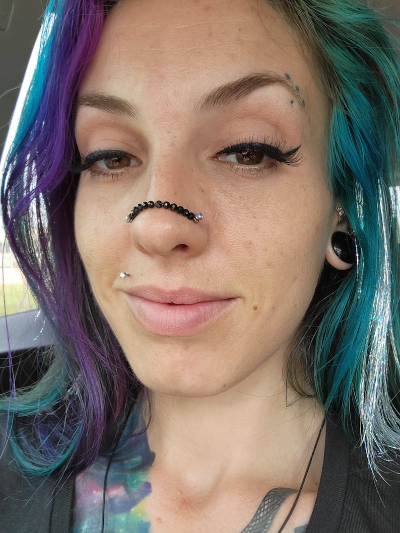 May include: A close-up of a person's face featuring a nose adorned with a black beaded nose chain. The person has multi-colored hair, with shades of blue, purple, and silver. The image also shows a lip piercing and a nose piercing.