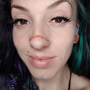 K&ouml;nnte beinhalten: Nahaufnahme eines Gesichts mit einem Septum-Piercing. Das Piercing ist ein Ring mit kleinen, orangefarbenen Perlen. Die Person hat blaue und gr&uuml;ne Haare und tr&auml;gt Eyeliner.
