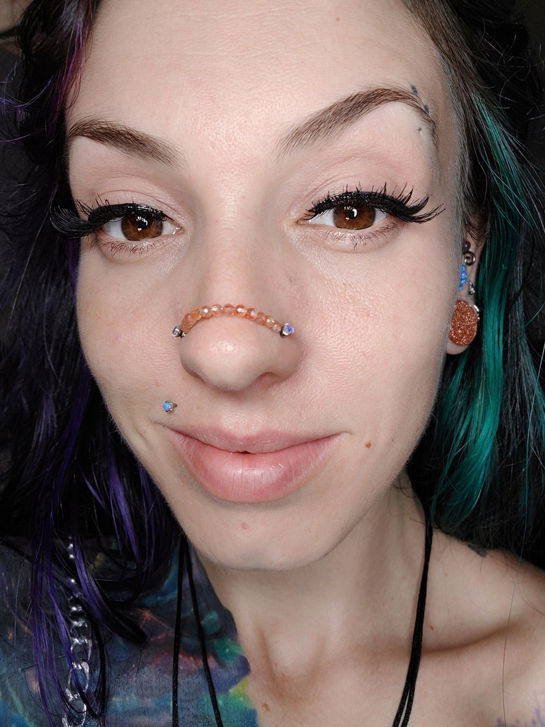K&ouml;nnte beinhalten: Nahaufnahme eines Gesichts mit einem Septum-Piercing. Das Piercing ist ein Ring mit kleinen, orangefarbenen Perlen. Die Person hat blaue und gr&uuml;ne Haare und tr&auml;gt Eyeliner.