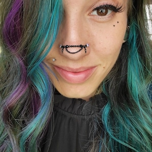 May include: A close-up of a person's face featuring a black chain nose ring with dangling chains and clear gems. The person has multi-colored hair with shades of teal, purple, and gray. The person is wearing a black top.