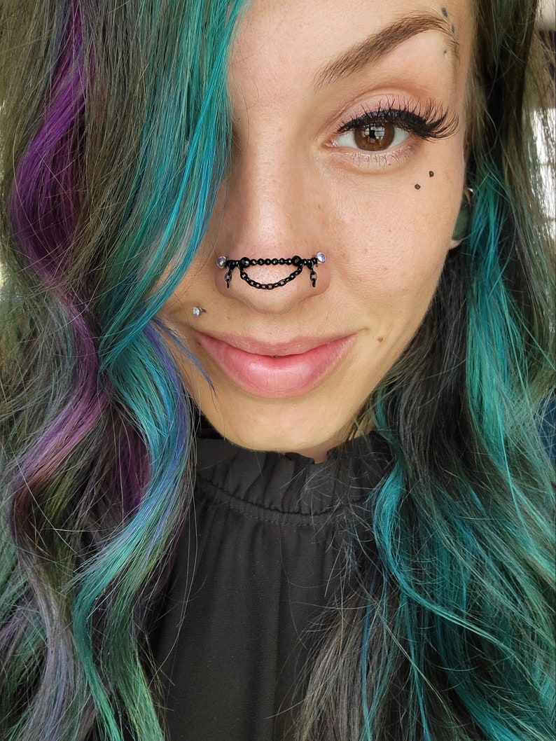 May include: A close-up of a person's face featuring a black chain nose ring with dangling chains and clear gems. The person has multi-colored hair with shades of teal, purple, and gray. The person is wearing a black top.