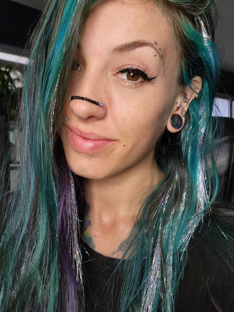 May include: A person with teal and purple hair, adorned with silver tinsel strands. They wear a black beaded nose chain and have multiple ear piercings with decorative jewelry. A large black ear gauge is visible.