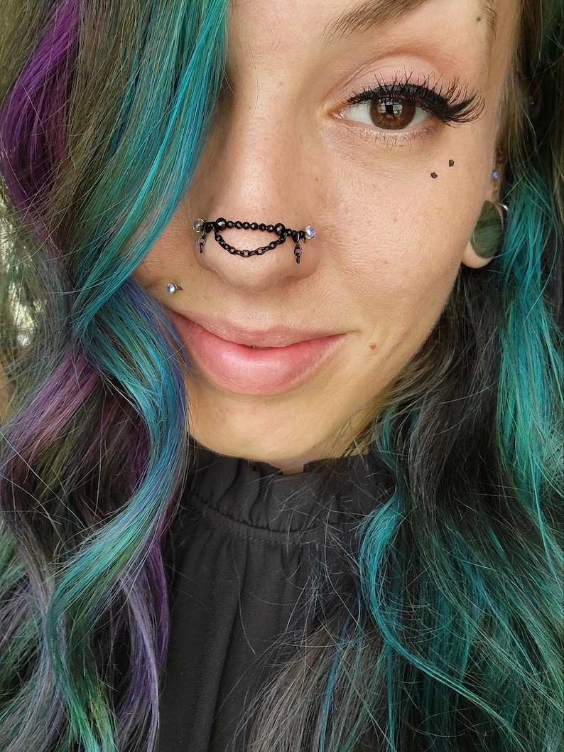 May include: Close-up of a person's face featuring a black nose chain piercing with dangling charms. The person has teal, purple, and green hair. The person is wearing a black top. The image is well-lit and in focus.