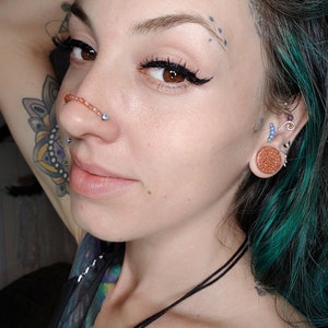 K&ouml;nnte beinhalten: Eine Frau mit t&uuml;rkisfarbenem Haar und mehreren Piercings. Sie tr&auml;gt einen Septumring mit einem kleinen blauen Stein, einen Nasenring und einen gro&szlig;en braunen Ohrstecker im rechten Ohr. Sie tr&auml;gt eine schwarze Halskette und eine silberne Kette.