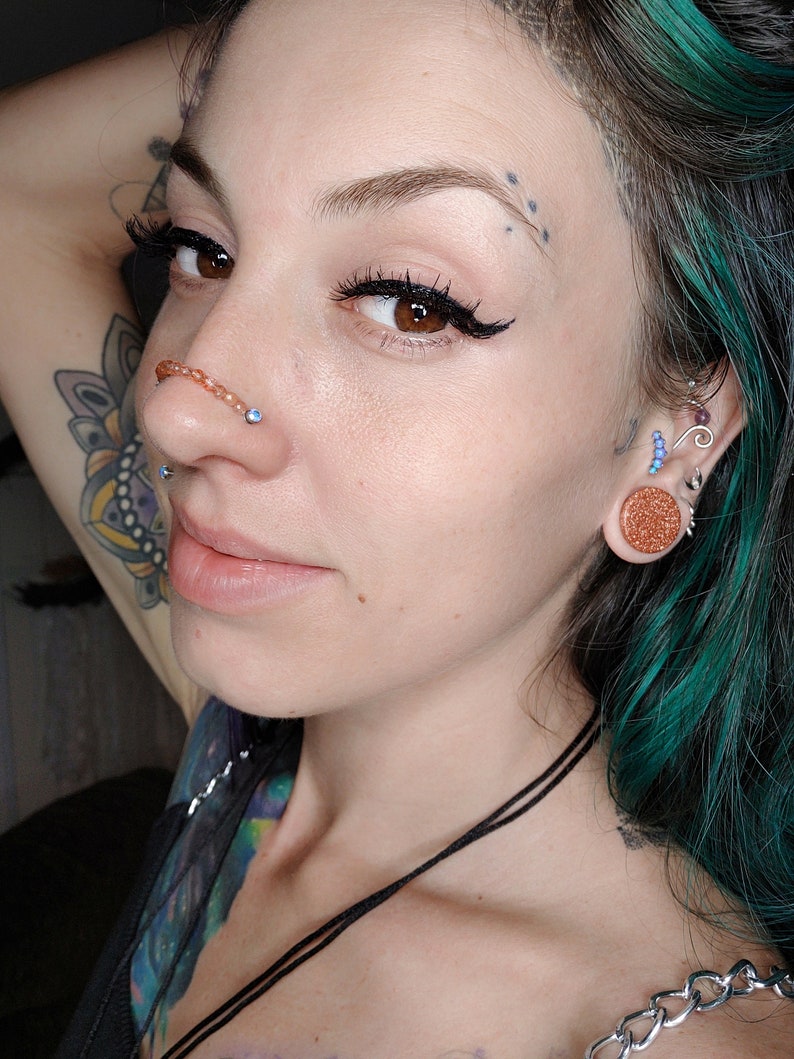 K&ouml;nnte beinhalten: Eine Frau mit t&uuml;rkisfarbenem Haar und mehreren Piercings. Sie tr&auml;gt einen Septumring mit einem kleinen blauen Stein, einen Nasenring und einen gro&szlig;en braunen Ohrstecker im rechten Ohr. Sie tr&auml;gt eine schwarze Halskette und eine silberne Kette.