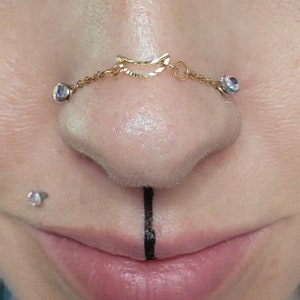 May include: A close-up of a person's face with a gold nose ring chain with two clear gems. The chain is attached to a small gold crescent moon shaped nose ring. The person also has a small black line drawn on their lip.