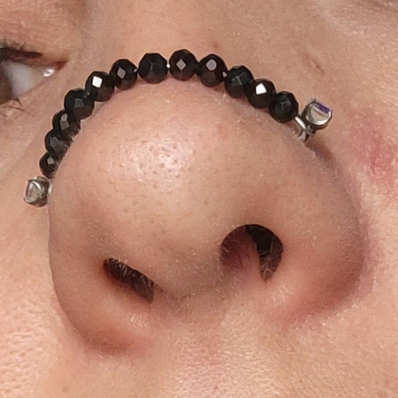 May include: A nose ring featuring a curved bar with a row of faceted black beads. The ends of the bar have small, silver-colored decorative elements. The jewelry is worn on the nose.