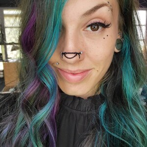May include: A close-up of a person with teal, purple, and gray hair. They are wearing a black nose chain with dangling accents and a black top. The person has multiple facial piercings and is smiling.