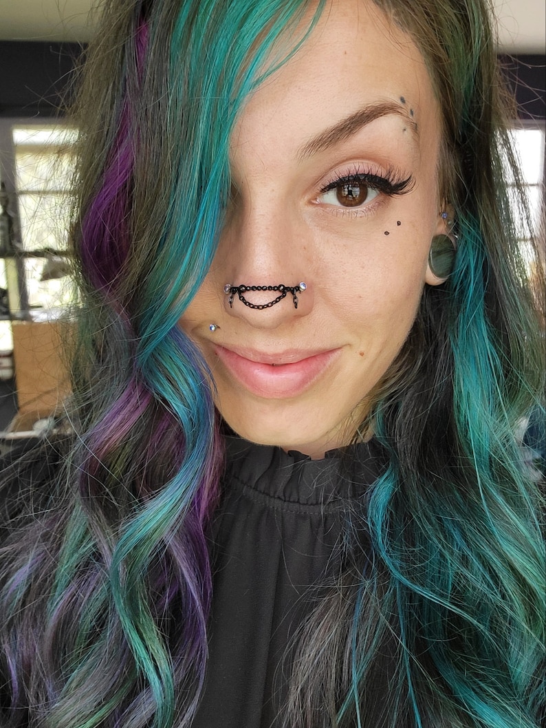 May include: A close-up of a person with teal, purple, and gray hair. They are wearing a black nose chain with dangling accents and a black top. The person has multiple facial piercings and is smiling.