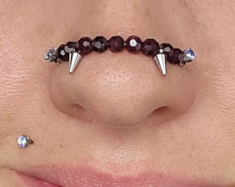 May include: A close-up of a person's nose with a septum piercing. The piercing is a row of small, dark purple beads with silver spikes on either end.