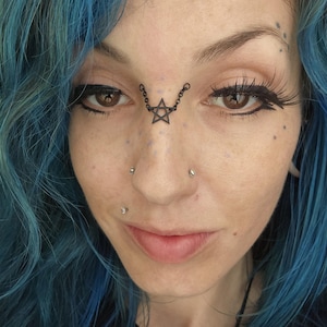 May include: A close-up of a person with blue hair and a black star-shaped face chain. The chain is connected to the eyebrows and nose. The person has multiple facial piercings and dark eyeliner.