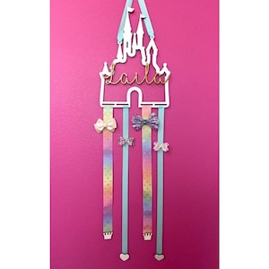 Puede incluir: Un soporte para accesorios para el cabello en forma de castillo blanco con el nombre "Laila" en letras doradas. El soporte tiene cuatro cintas colgantes con patrones de arcoíris y coronas blancas en la parte inferior. También hay varios lazos coloridos unidos a las cintas.
