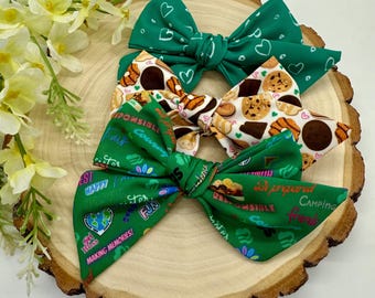 May include: Three green fabric hair bows with different designs. One bow has a green heart pattern, one has a cookie pattern, and one has a green pattern with text that says "I'm prepared", "responsible", "courage is", "independence", "fun", "happy", "helpful", "sister", "best", "making memories", and "camping with friends".