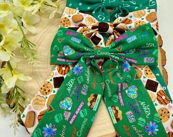May include: Green and white fabric bow with a Girl Scout theme. The bow features a variety of Girl Scout cookie designs, including Thin Mints, Samoas, and Tagalongs. The bow also features the Girl Scout logo and the words "Camping", "Sister", "Fun", and "Confidence".