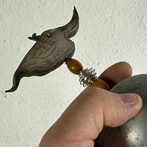 Könnte beinhalten: Eine Metallskulptur mit einer grauen Kugel und einem rechteckigen Sockel. Die Skulptur hat ein dekoratives Element mit einem geschnitzten Design und einer Feder. Die Skulptur wird in einer Hand gehalten.