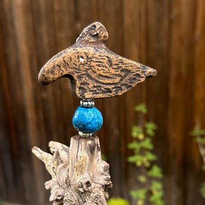 Könnte beinhalten: Handgefertigte Holzskulptur mit einem Vogel auf einem Treibholzsockel. Eine leuchtend blaue Perle akzentuiert das Design. Die Skulptur ist auf einem quadratischen, cremefarbenen Pfosten montiert, der natürliche Texturen und Farben zeigt.