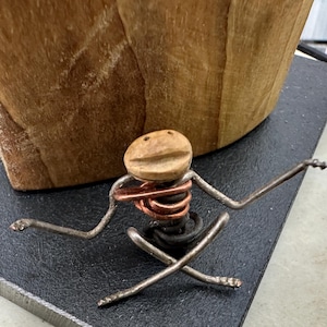 Könnte beinhalten: Eine Metallskulptur einer Person in einer sitzenden Yoga-Pose. Die Figur besteht aus silberfarbenem Draht, Kupferdraht und einem Holzkopf. Die Skulptur befindet sich auf einer dunkelgrauen quadratischen Oberfläche, mit einem großen Holzobjekt im Hintergrund.