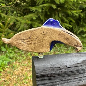 Puede incluir: Una escultura de pez de cerámica hecha a mano con un cuerpo marrón y detalles azules. El pez tiene un ojo pequeño y redondo y una cuenta decorativa adjunta en la parte inferior. La escultura se exhibe al aire libre sobre un fondo de follaje verde.