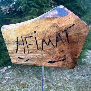 Könnte beinhalten: Ein Holzschild mit dem Wort "HEIMAT" in schwarzen Buchstaben geschnitzt. Das Schild ist hellbraun mit einer dunkelbraunen Oberseite und einem kleinen Blumendesign. Es ist auf einer Metallstange montiert.
