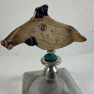 May include: A distinctive ceramic sculpture with a tan, wing-shaped design, dark blue details, and a spiral motif. It is mounted on a silver-toned base, with teal and silver bead accents. The sculpture is a unique piece of art.