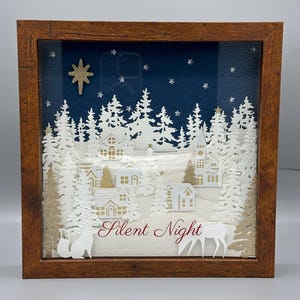 Pode incluir: Uma caixa de sombra de Natal emoldurada com uma cena de vila de inverno. A obra de arte inclui recortes de papel branco de casas, árvores e um veado, contra um fundo de céu estrelado azul escuro. As palavras "Silent Night" estão escritas em vermelho.