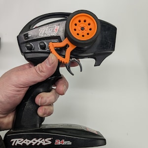 Peut inclure: Une télécommande noire et orange pour une voiture jouet. La télécommande a un volant, une gâchette et plusieurs boutons. La télécommande est étiquetée "Traxxas 2.4 GHz".