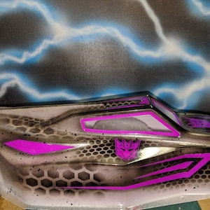 May include: A black and grey car body with a honeycomb pattern and purple accents. The car body has a purple Transformers logo on the side.